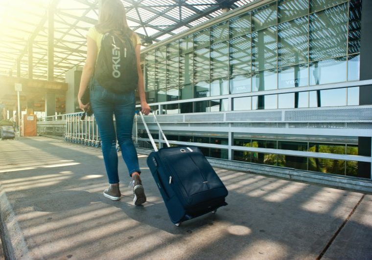         A woman with a backpack is walking with a suitcase in front of an airport, possibly leaving her home.